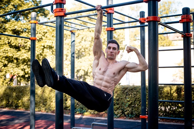 Strong man hanging from a pull-up bar at an outdoor workout park, flexing his biceps and showing his abs during a home weight loss workout.