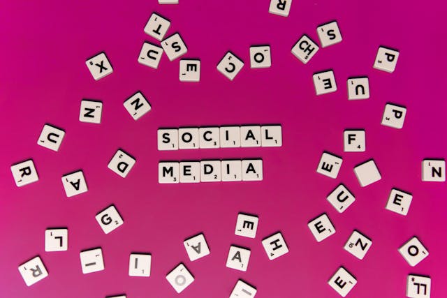Scrabble tiles on a pink background spelling out the words "SOCIAL MEDIA," surrounded by scattered letters. This image represents the strategic thinking needed to determine the best social media posting frequency for a business. 